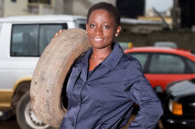 Young mechanic standing and smiling has a tire on her shoulder in her studio. clipart
