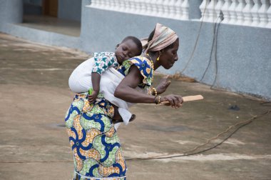 Anekro, Ivory Coast - August 21, 2015: old woman dancing with a child on the back. A woman dressed in loincloth, head tied with a scarf dance and lowering a child on her back clipart