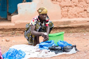 Abican, Fildişi Sahili - 22 Şubat 2017: çok renkli bir eşarp tarafından bağlı ve manyok un, içeren bir tepsi tutarak başını bir taburede oturan Afrikalı kadın kameraya benziyor. Tam attit, tüm Ivorians tarafından sevilen gıda üretimini mevcuttur