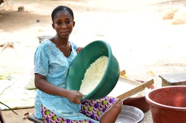Abidjan, cte Sahili - 22 Şubat 2017: çok renkli bir eşarp tarafından bağlı ve manyok un, içeren bir tepsi tutarak başını bir taburede oturan Afrikalı kadın kameraya benziyor. Tam attit, tüm Ivorians tarafından sevilen gıda üretimini mevcuttur