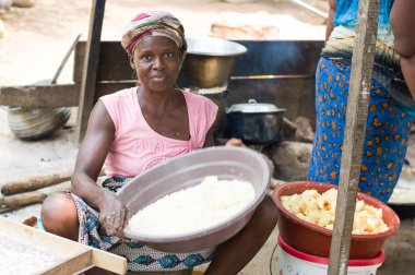 Abidjan, cte Sahili - 22 Şubat 2017: çok renkli bir eşarp tarafından bağlı ve manyok un, içeren bir tepsi tutarak başını bir taburede oturan Afrikalı kadın kameraya benziyor. Tam attit, tüm Ivorians tarafından sevilen gıda üretimini mevcuttur