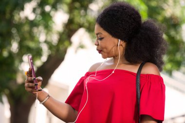 Kırmızı tişörtlü, kulaklıklı mutlu genç bir kadın şehirde cep telefonuyla konuşuyor.