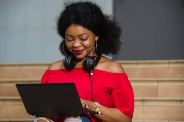 Boynunda kulaklıkla oturan genç kız dizüstü bilgisayara bakıp gülümsüyor..