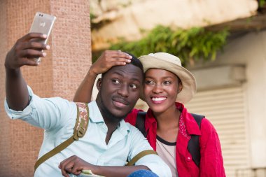 Bir çift genç turist dışarıda oturmuş gülümserken kendilerini cep telefonuyla fotoğraflıyorlar..