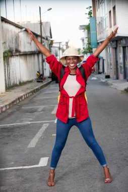 Sokakta pantolonlu, sırt çantalı genç bir kız kameraya bakıp gülümsüyor..