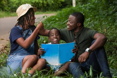 Çimlerin üzerinde oturan bir çift birbirlerine bakıp gülümsüyorlar ve yanlarında oğulları var..