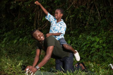Parkta babasının sırtında oturan genç bir çocuk yumruğunu kaldırmış gülüyor..