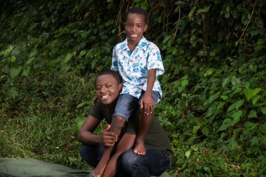 Genç bir adam parkta çömelmiş oğlunu sırtında taşıyarak kameraya bakıp gülümsüyor..