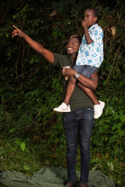 Omzunda çocuğuyla parkta duran genç bir adam ona gülümseyen bir şey gösteriyor..