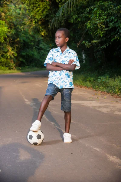 a young boy standing in a park with a ball under his foot and looking in profile with his arms crossed.