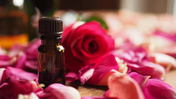 Bottle of aroma oil with red rose and roses petals on wooden surface, selected focus