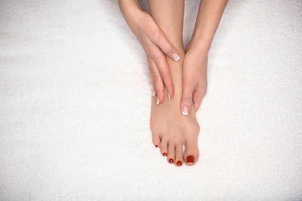 Female foot on white terry towel and hands. Woman holds her sole and mass it by her fingers. Classic French manicure and red pedicure. — Stock Photo, Image