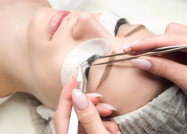 Eyelash extension procedure. Woman face and experts fingers with tweezers, close up, selective focus — Stock Photo, Image