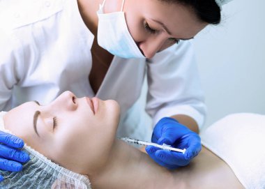Güzellik uzmanı, hasta yüzü ve elinde tek kullanımlık şırınga ile tek tip kıyafetli güzellik salonunda çalışıyor. Müşteri kapalı gözleri ile bir kanepede yatıyor ve prosedür için hazır.