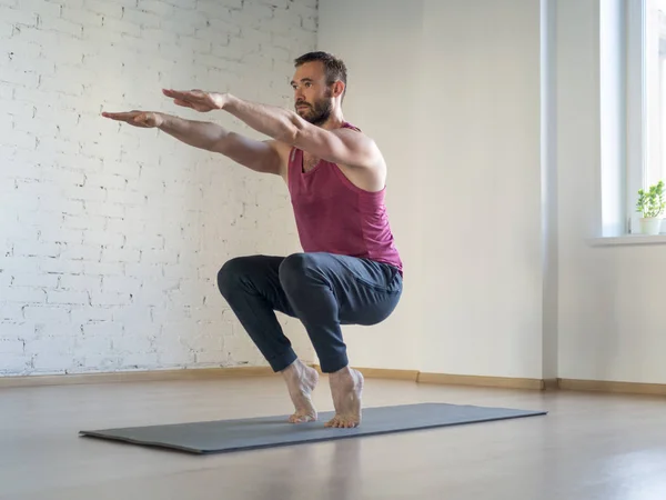 Caucasian man stands on tiptoe and practice yoga in fitness studio, loft style, selective focus. Chair pose.