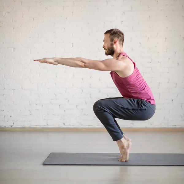 Chair pose. Caucasian man stands on tiptoe and doing yoga in fitness studio, side view, selective focus.