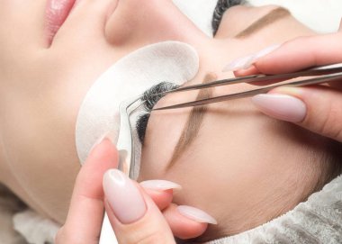 Kirpik uzatma işlemi. Uzun siyah kirpikli kadın yüzü ve cımbızlı usta parmakları, yakın çekim, seçici odak.