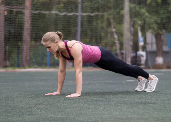 Blond dziewczyna robi deski ćwiczenia na boisko sportowe na zewnątrz, selektywne skupienie — Zdjęcie stockowe