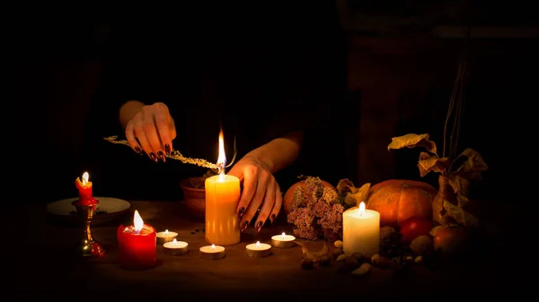 Strega fare un incantesimo sull'altare al buio. Mani femminili con unghie nere taglienti che bruciano erba magica tra candele, zucca, noci, foglie secche, messa a fuoco selezionata, basso profilo — Foto Stock