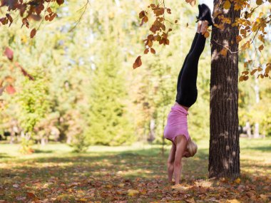 Atletik kadın, sonbaharda şehir parkındaki ağacın yanında klasik bir amuda kalkıyor.