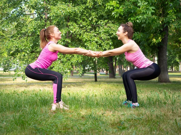 Żeński trening w parach na świeżym powietrzu. Dwie białe śmiejące się dziewczyny robią przysiady i pomagają sobie nawzajem, w parku publicznym na trawie w lecie, selektywne skupienie — Zdjęcie stockowe