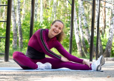 Gülümseyen, beyaz, mavi spor giysili güzel bir kadın spor salonunda, spor alanında, yazın, seçici bir şekilde bacaklarını esneterek oturuyor..