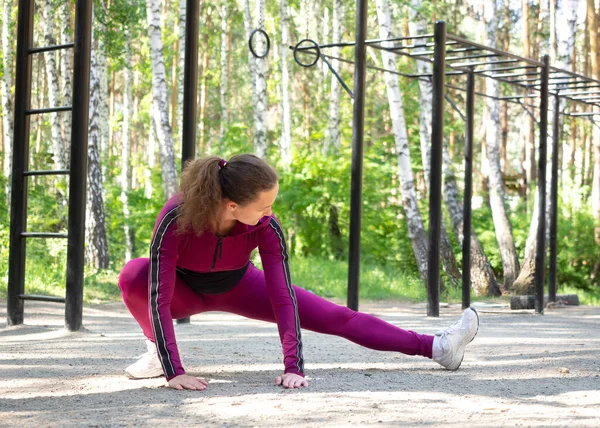 Tipikus bemelegítés edzés előtt. Fitt kaukázusi lány bíboros sportruházat nem lábak nyújtózik a földön a sport területén szabadtéri nyáron, szelektív fókusz. — Stock Fotó