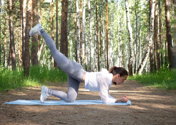 Pasująca biała dziewczyna w białej odzieży sportowej ćwiczy jedną nogę. Trening na świeżym powietrzu w parku, w lecie, selektywne skupienie. — Zdjęcie stockowe