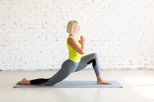 Hip joint mobility. Fit adult caucasian woman practice stretching in deep lunge on a mat in loft white studio indoor, selective focus.