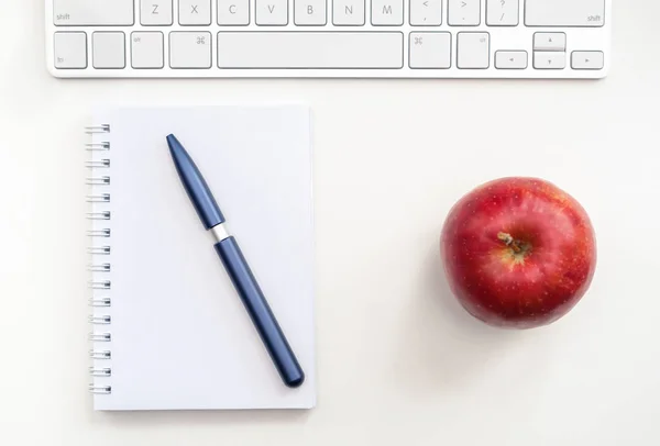 A fragment of the workplace with a keyboard, an opened notebook and a red apple on a white desk