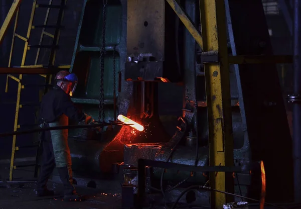 Hot metal ingot being loaded into a hammer forge. Worker forges iron products