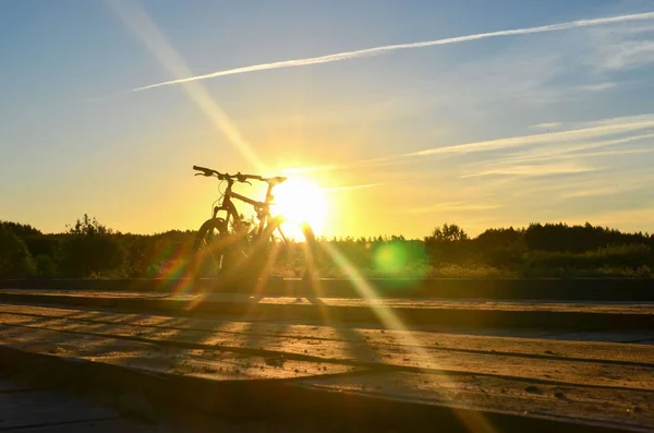 Bright sunrise on the road near the river on the background of a bicycle. Mountain bike in forest with sun rays. Walking along the shore with the bike against sunset sky with clouds and sun rays.