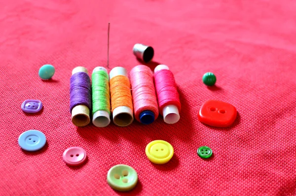 Items for sewing clothes, colorful spools of thread, buttons, needle and thimble against the background of red fabric. Needlework and tailoring concept