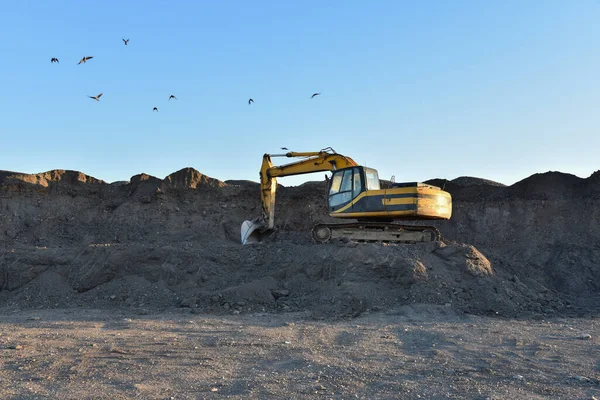 Excavator working at open pit mining. Backhoe digs gravel in sand quarry on blue sky background. Recycling old asphalt at a landfill for the disposal of construction waste