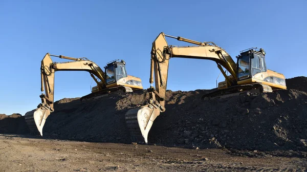 Excavators working at construction site. Backhoe digs ground in sand quarry on blue sky background. Construction machinery for excavation, loading, lifting and hauling of cargo on job sites