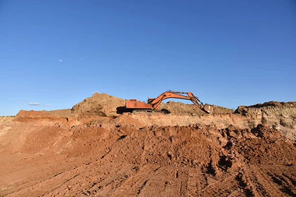 Excavator working at construction site. Backhoe digs ground in sand quarry on blue sky background. Construction machinery for excavation, loading, lifting and hauling of cargo on job sites