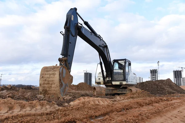 Black and white excavator during earthworks at construction site. Backhoe digging the ground for the foundation and for laying sewer pipes district heating. Earth-moving heavy equipment