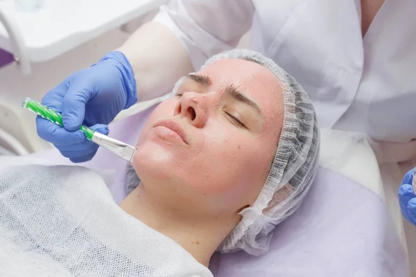 Crop master applying white mask with brush on face and neck of young woman in towel lying on table — Stock Photo, Image