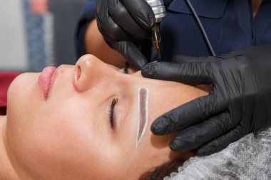 Usta, bir dövme makinesiyle müşteriye kaş makyajı yapar.