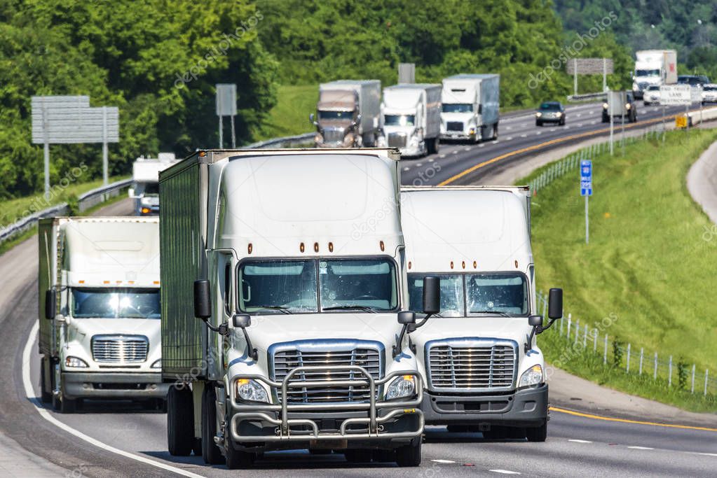 A constant line of heavy trucks move down a busy interstate.  Note: All logos and identifying marks have been removed from all vehicles.  Image was created on hot day, so heat waves from the asphalt create some distortion, especially on vehicles fart