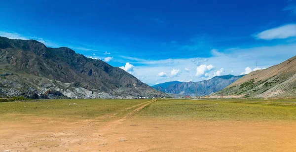 Mountain valley with a village far away The Republic of Altai, Russia. A field with a dirt road in the foreground, large mountains, far away a village and a small part of an asphalt road