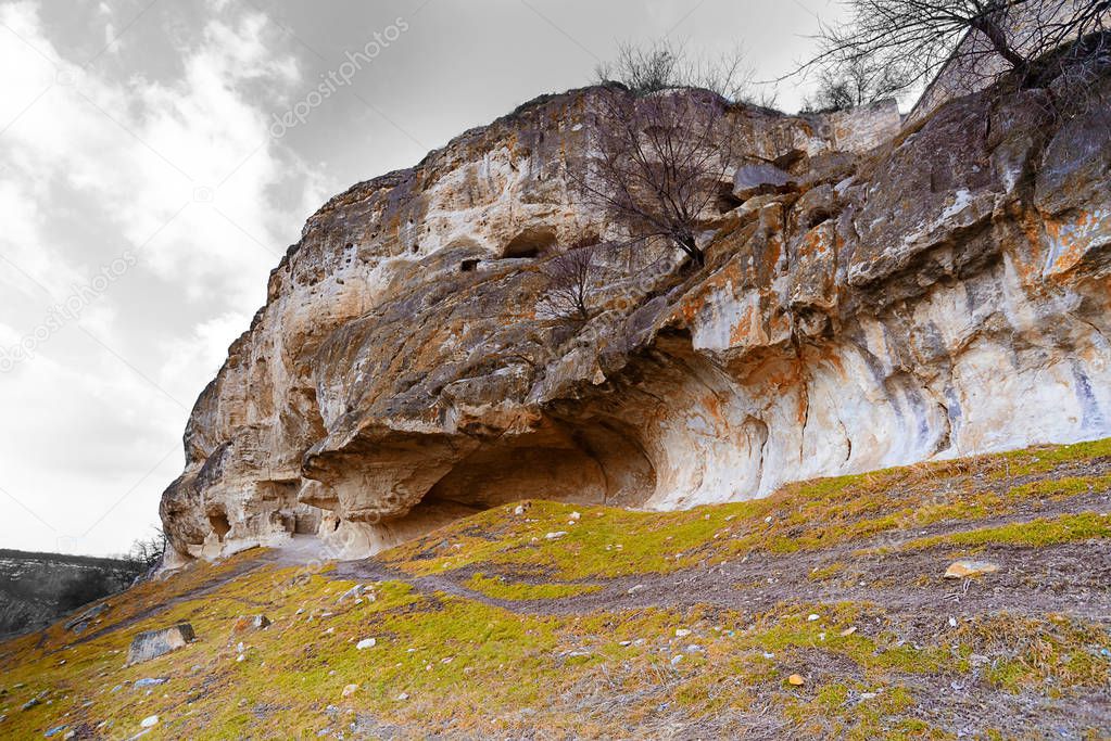 Cave city Chufut-Kale, view from the bottom of the mountain, Bakhchisaray district, Crimea, Ukraine. Foreground - fragments of snow, golden grass, the far plan - cave mountain, gray sky