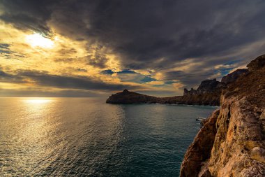 Denizdeki günbatımının panoramik görüntüsü, Cape Kapchik, Golitsyna Patikası, Novy Svet, Sudak, Kırım, Ukrayna. Sağda, güneşte bir kaya parçası, bir deniz, uzakta uzun bir pelerin, parlak renkli bulutlar.