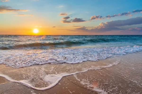 Zonsondergang met een zonneschijf aan de hemel met wolken en witte schuimgolven op het zand op de voorgrond. — Stockfoto