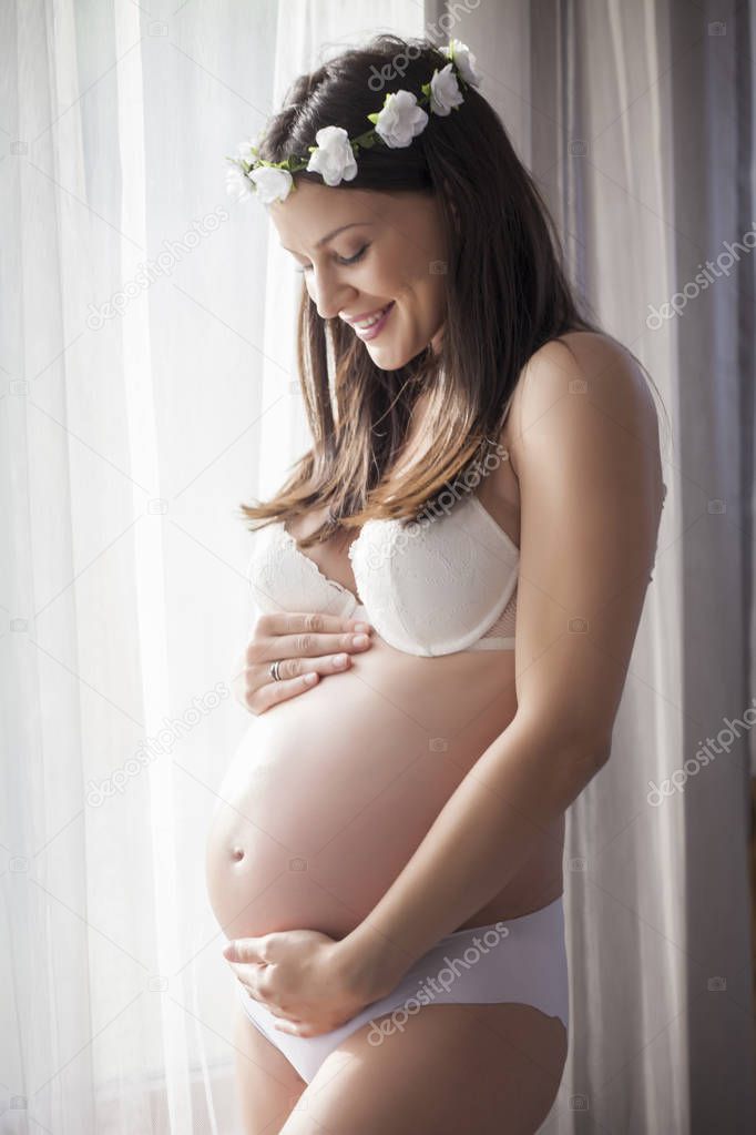 Happy pregnant woman in underwear by the window with curtains and a wreath of flowers on her head