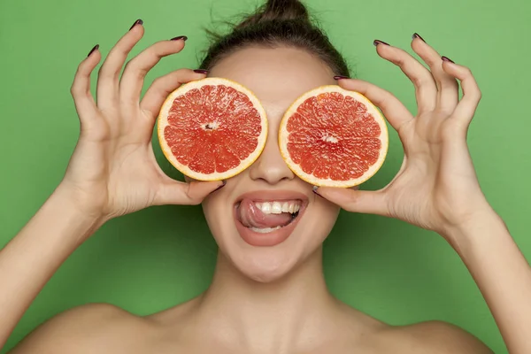 Sexy young woman posing with slices of red grapefruit on her face on a green background