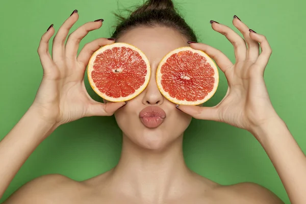 Sexy young woman posing with slices of red grapefruit on her face on a green background