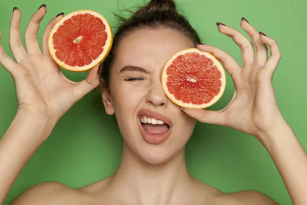 Funny young woman posing with slices of red grapefruit on her face on a green background