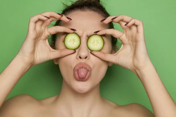 Happy sexy woman posing with slices of cucumber on her face on a green background