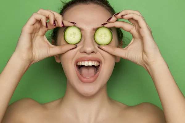 Happy young woman posing with slices of cucumber on her face on a green background